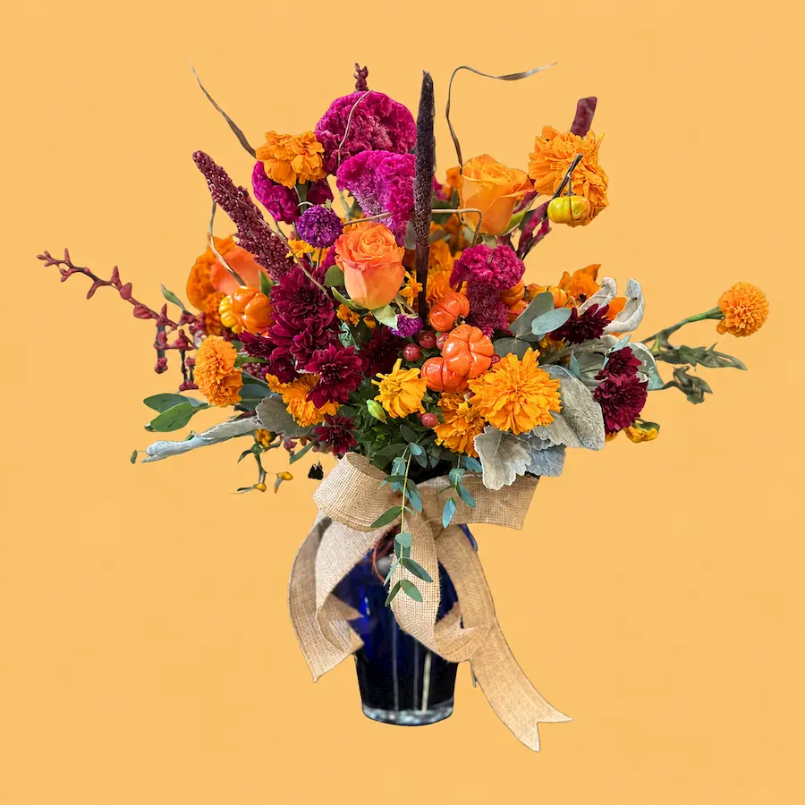 Arreglo Mictlán con cempasúchil naranja, garra de león magenta, rosas y calabazas mini en un cilindro de cristal con moño de yute.