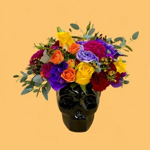 Mirada de Catrina, calavera de cerámica negra con flores naturales amarillas, naranjas, rojas, moradas y magenta, acompañadas de follaje verde.
