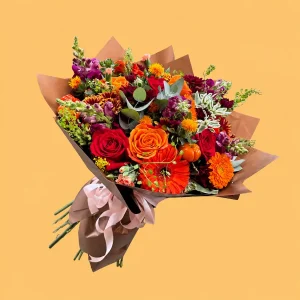 Ramo Tierra de Recuerdos con rosas rojas y naranjas, gerberas, perritos morados y calabazas mini, envuelto en papel café con listón rosa claro.